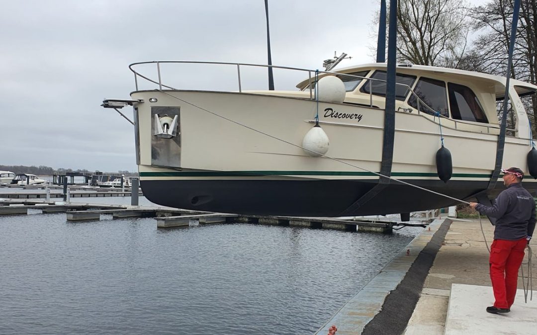 Cremefarbene Motoryacht in einem Hafen, am Kran aufgehängt, ein Bootsführer hält eine Halteleine